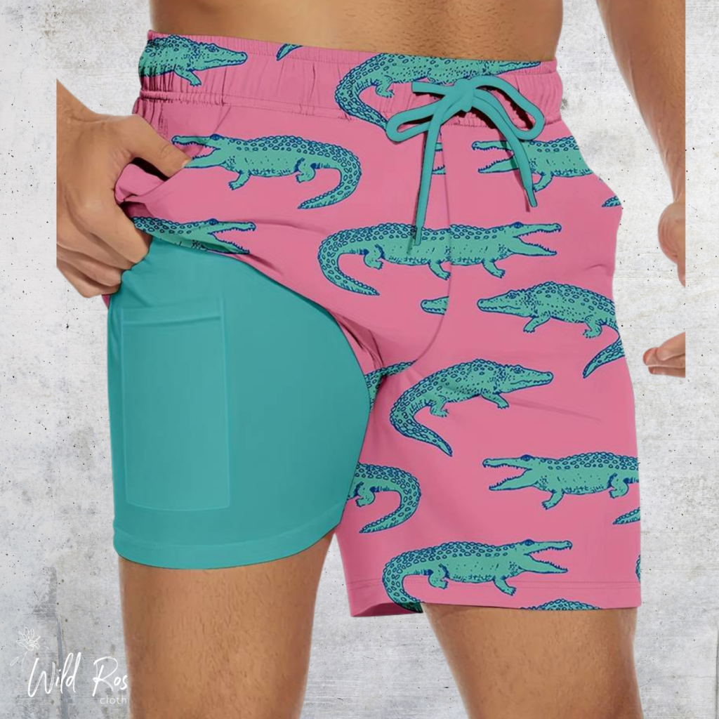 Croc Bay Swim Shorts featuring a playful teal crocodile print on a pink background, with contrasting teal inner shorts, an elasticated waistband and adjustable drawstring.
