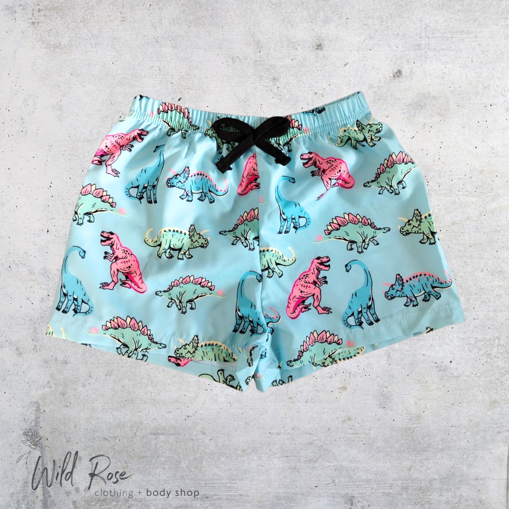 Dino Splash Swim Shorts – Kids Dinosaur Swimwear | WildRose Dino Splash Swim Shorts featuring colourful pink, blue, green and turquoise dinosaurs on a light blue background, perfect for kids' swimming and beach days.